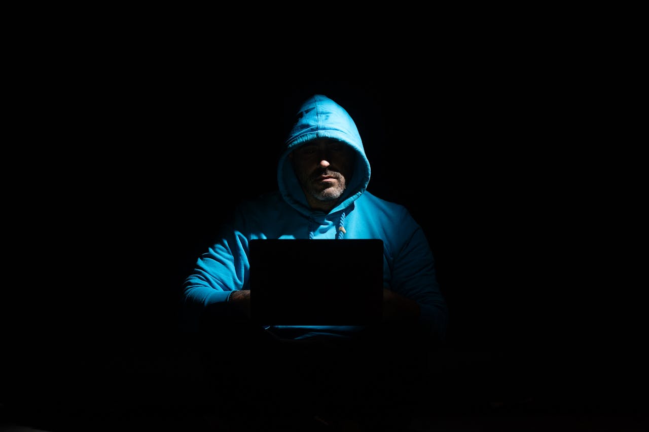 A person in a hoodie using a laptop in a dark setting, conveying mystery and technology themes.
