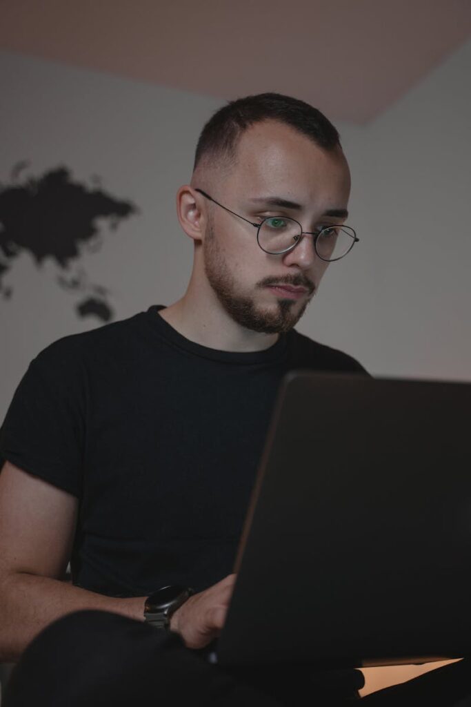 Man with eyeglasses and beard working on a laptop indoors, focused and engaged.